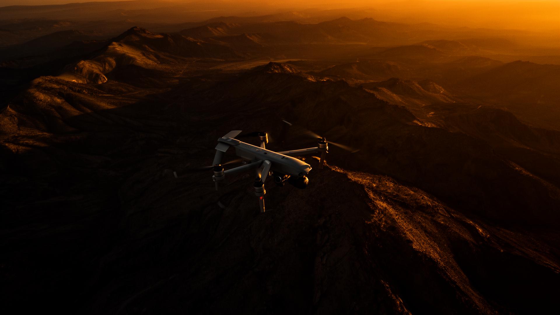 Unmanned aerial vehicle flying over amber-lit mountain terrain at dusk